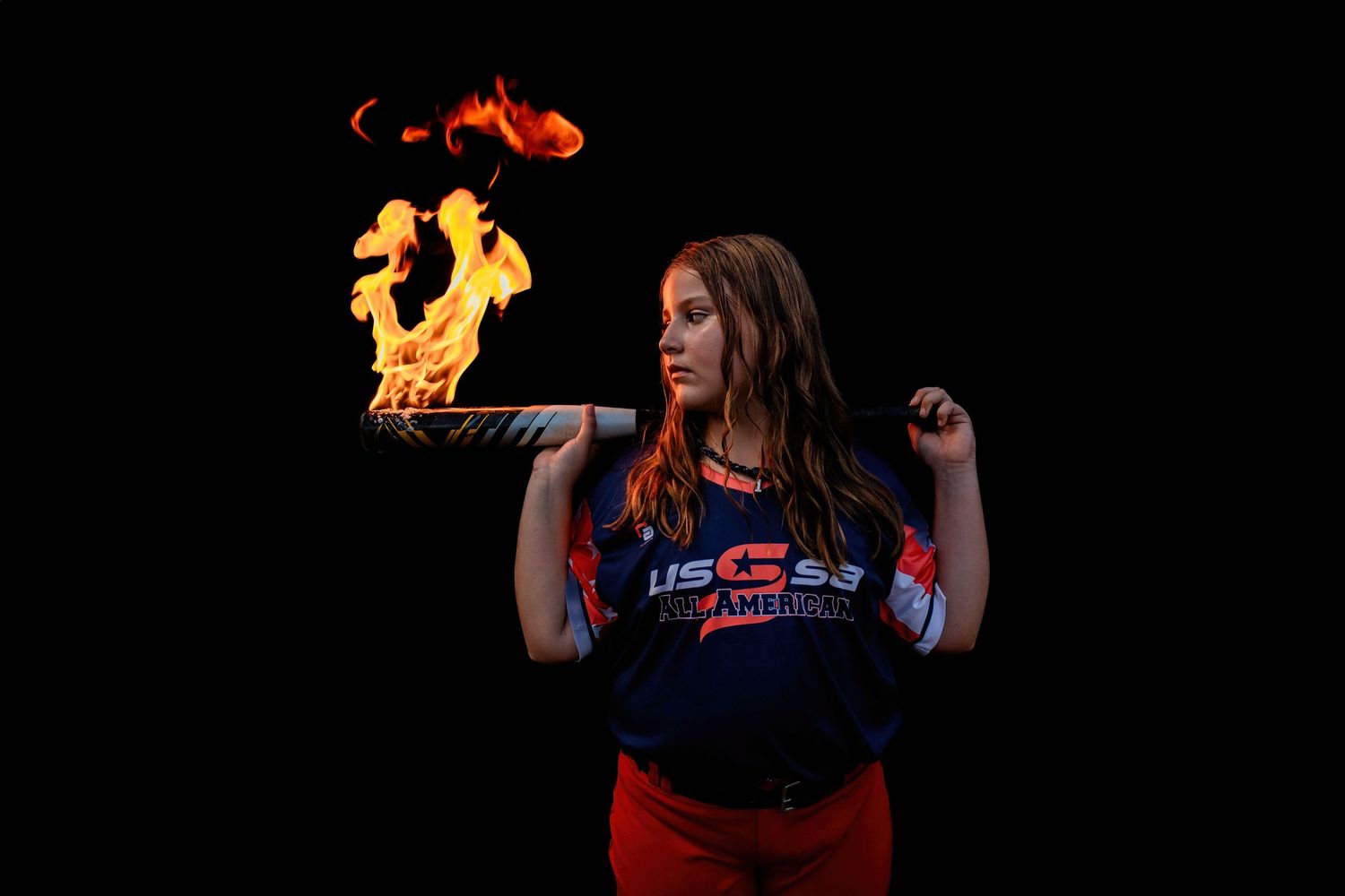 Athlete performs trick with flaming softball bat in dramatic dark setting.