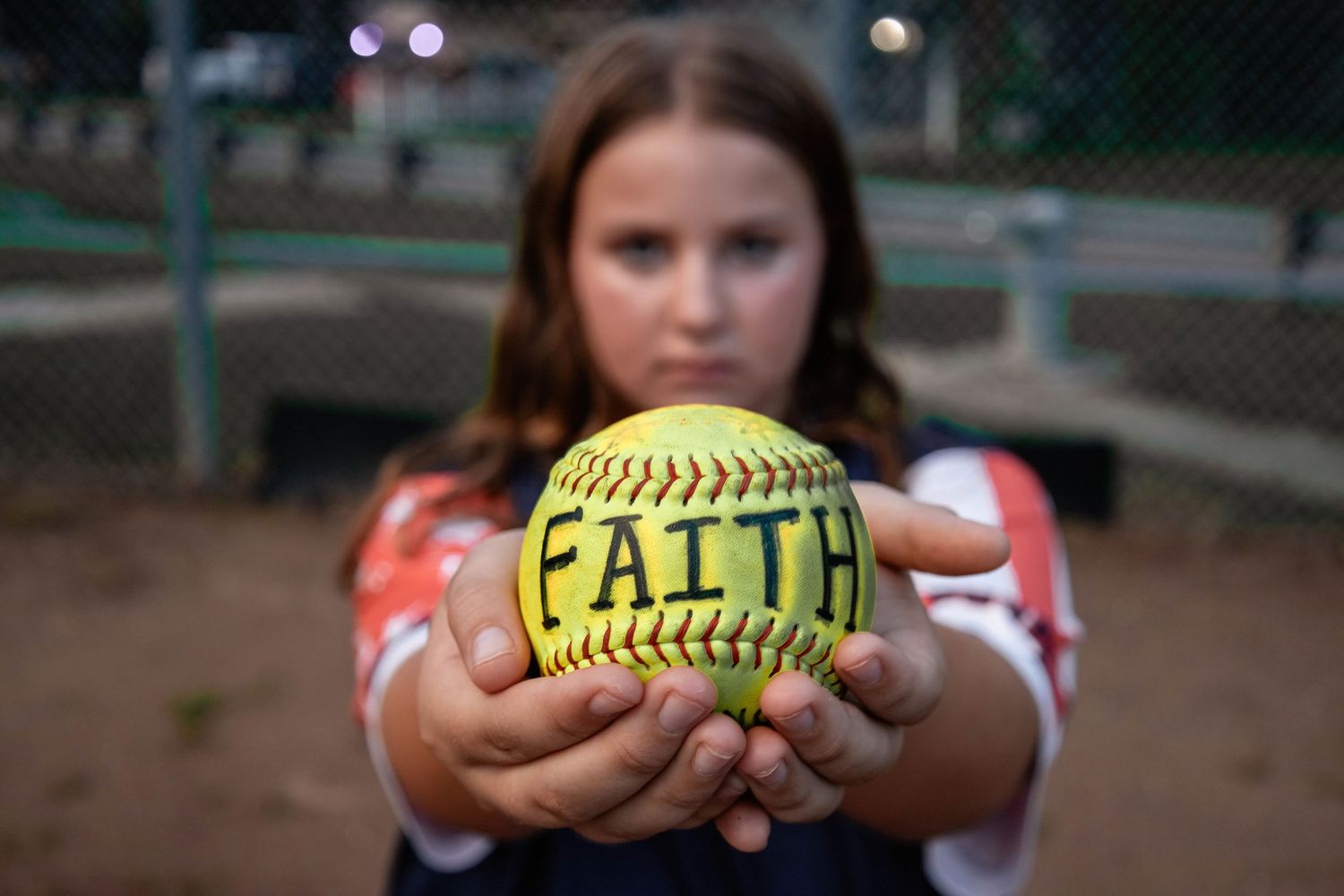 Softball player holds up yellow ball with FAITH written on it in close-up shot.