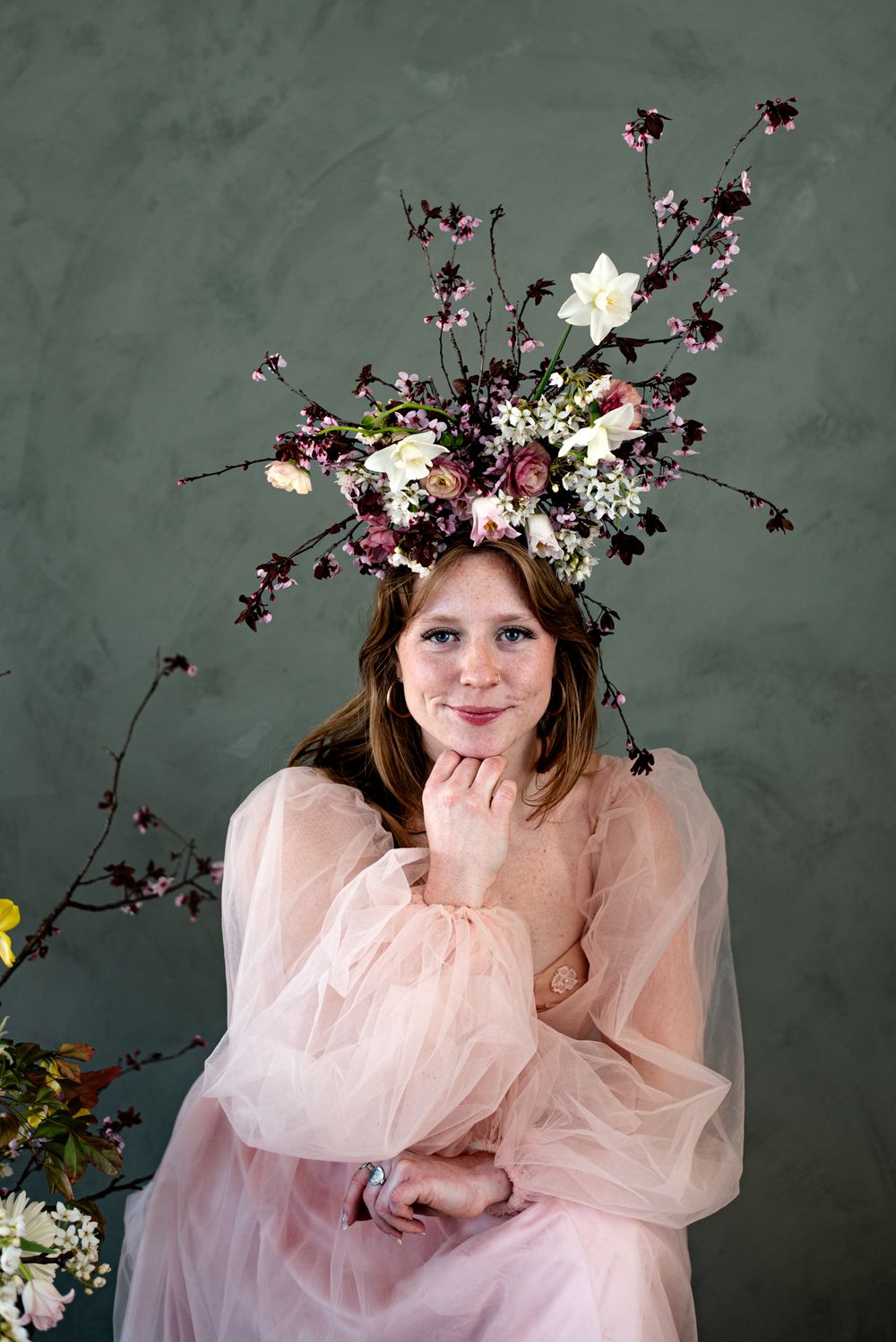An ethereal pink tulle dress adorned with a dramatic floral crown featuring white and burgundy blooms against a dark green wall.