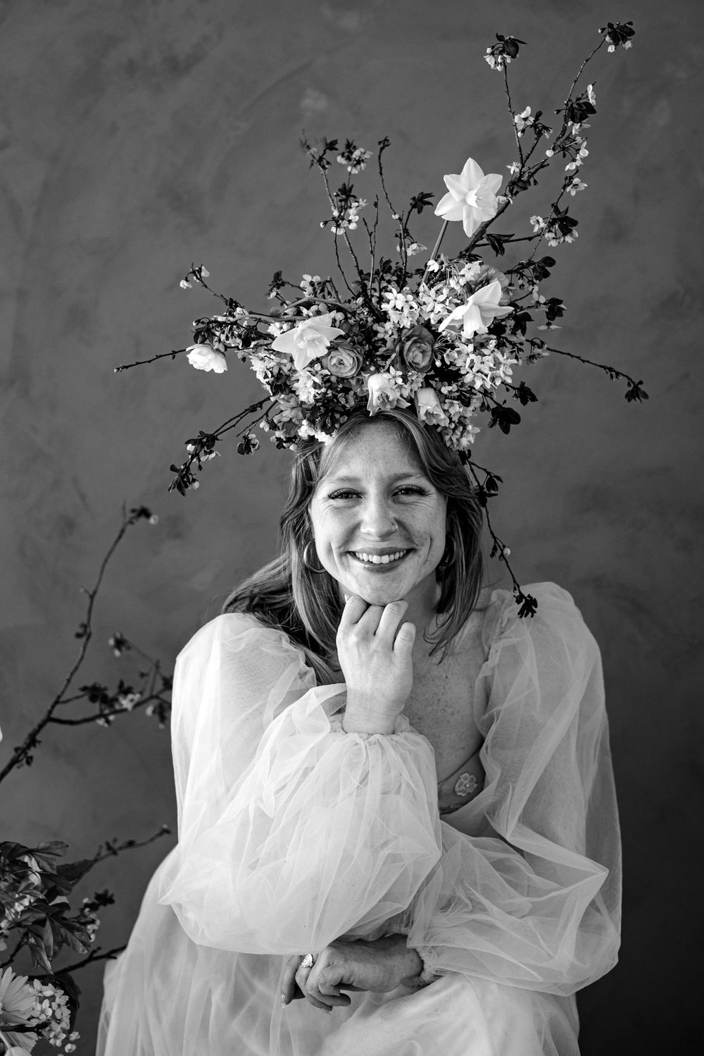 Black and white portrait showing someone in a flowing white dress adorned with a dramatic floral headpiece.