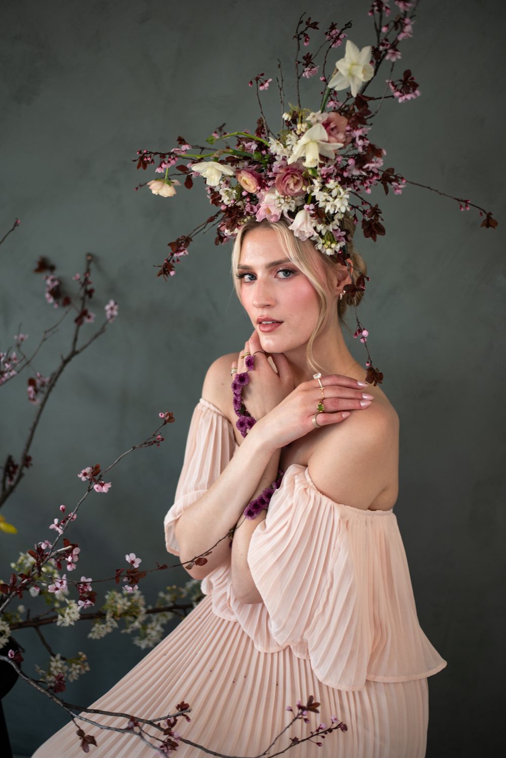 Ethereal spring portrait with pink pleated off-shoulder dress adorned with cherry blossoms and floral crown against gray backdrop.