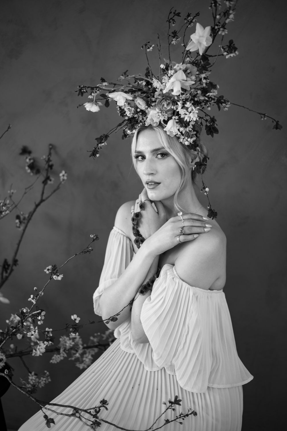 Black and white portrait with floral crown and flowing off-shoulder pleated dress surrounded by delicate branches.