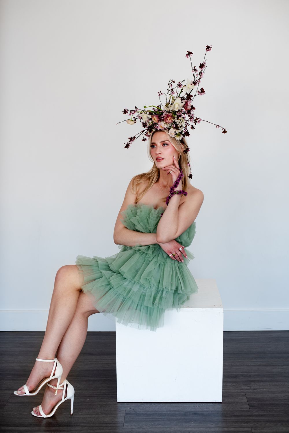A figure in a mint green tulle dress sits on white cube wearing strappy heels and floral headpiece against gray wall.