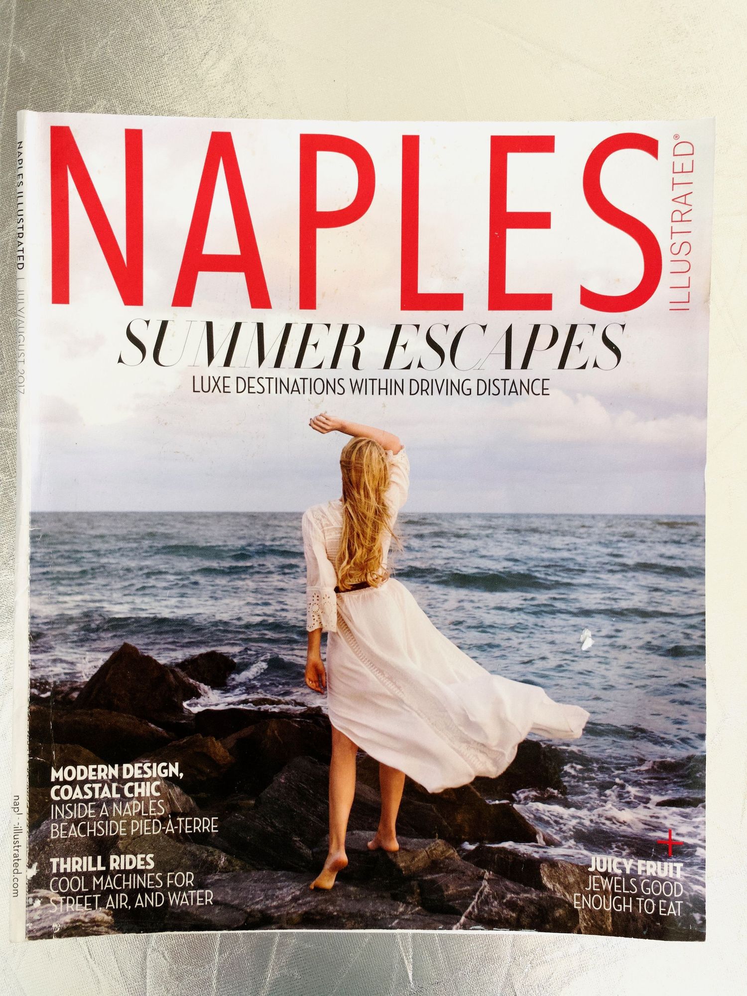 Naples Illustrated Summer Escapes magazine cover featuring woman in white dress on rocky coastal shore.