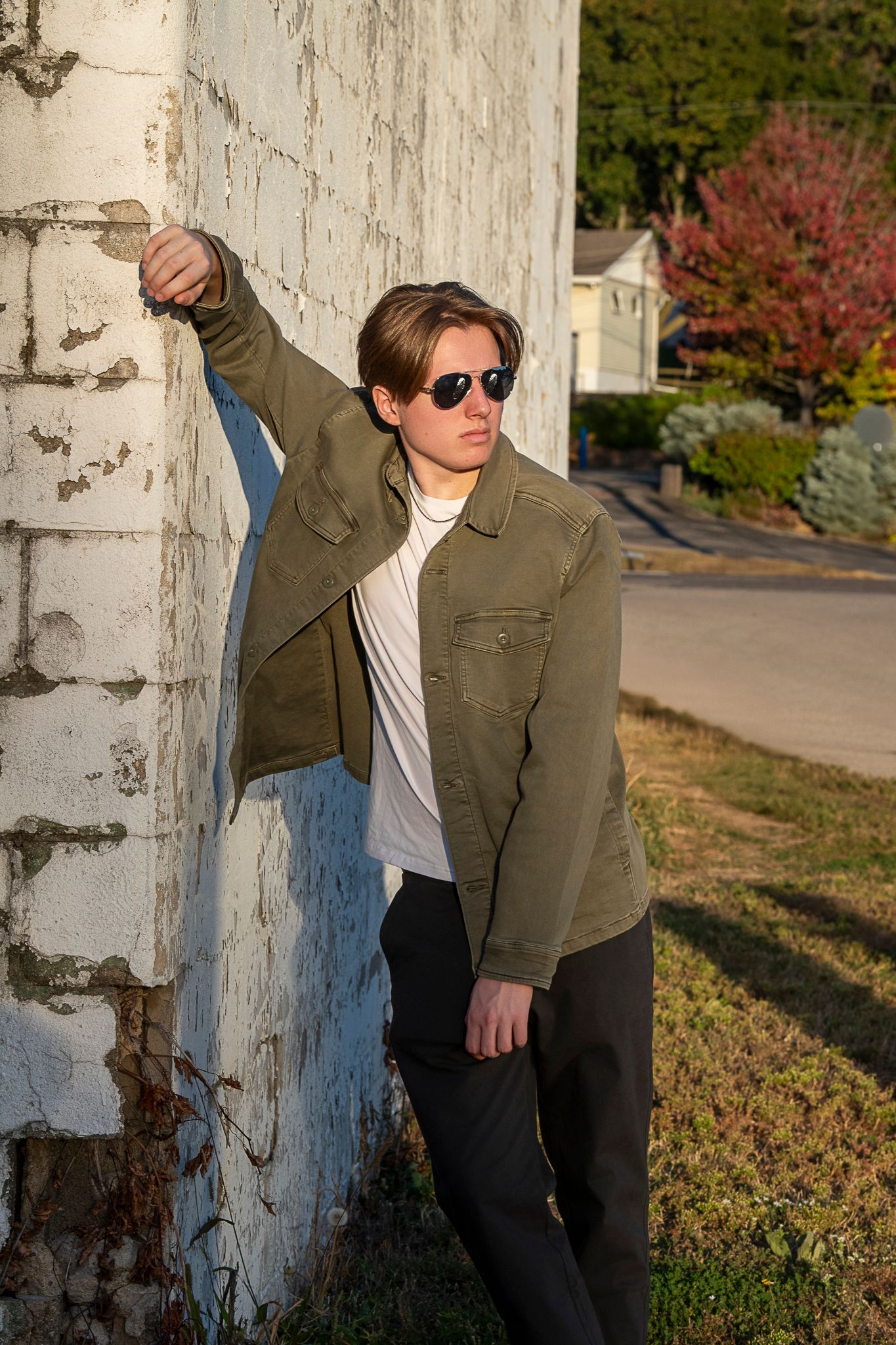 A person in a green jacket and sunglasses leans against a white wall during golden hour on an autumn day.