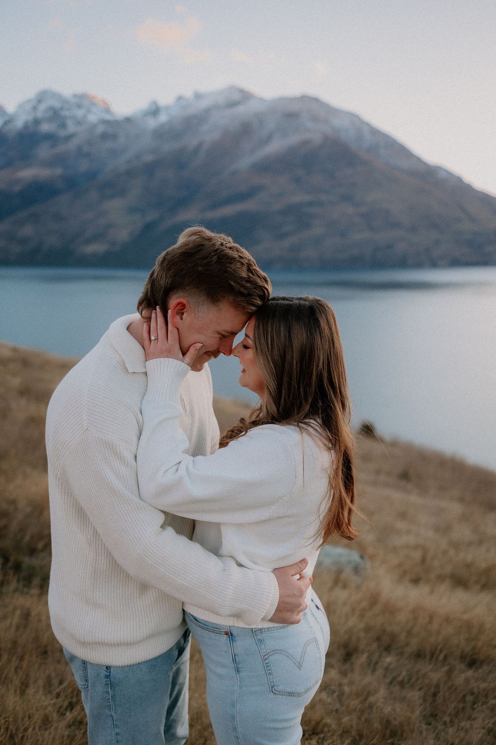 Couple in white sweaters and jeans embracing with foreheads touching, scenic mountain lake backdrop at dusk.