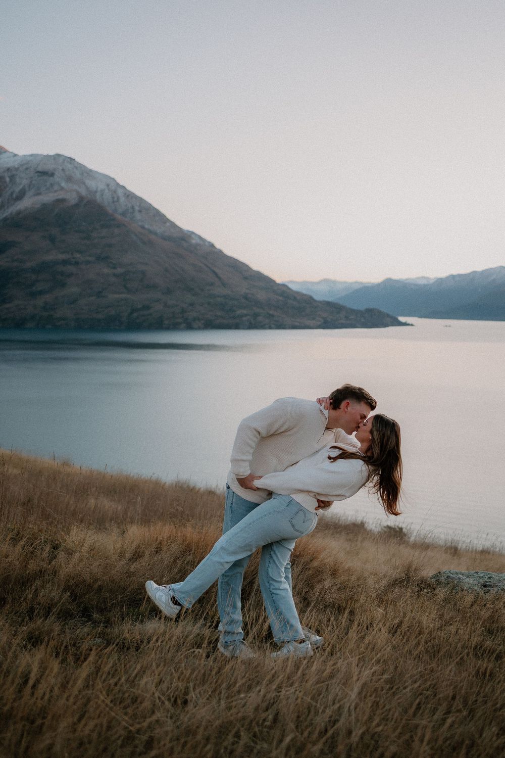 Couple sharing a romantic dip kiss on a hillside overlooking a serene mountain lake at dusk.