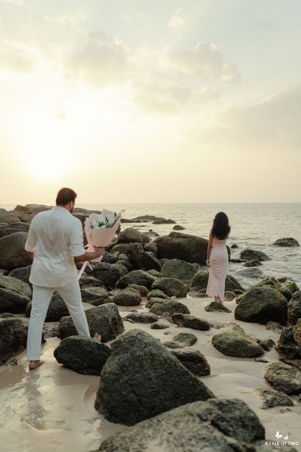 Romantic photoshoot on a rocky beach during golden hour with ocean waves in background.
