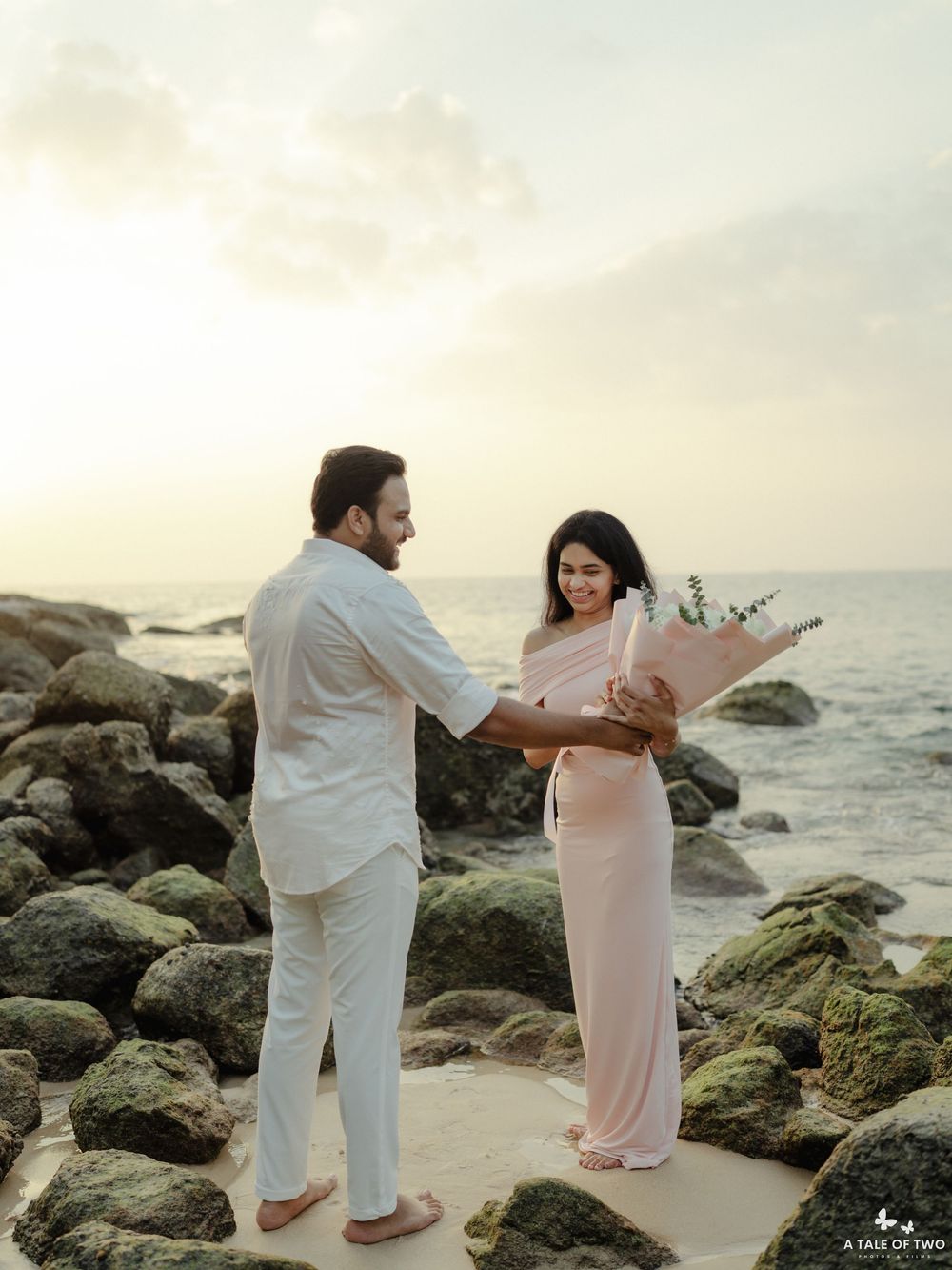 A couple in white and pink outfits poses on a rocky beach during sunset.