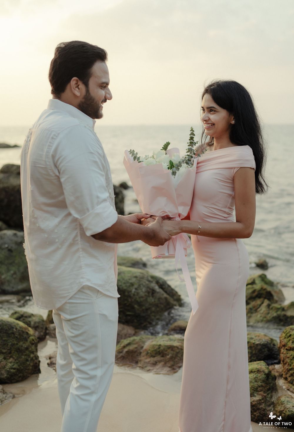 Couple sharing a romantic moment on a rocky beach with a pink bouquet at sunset.