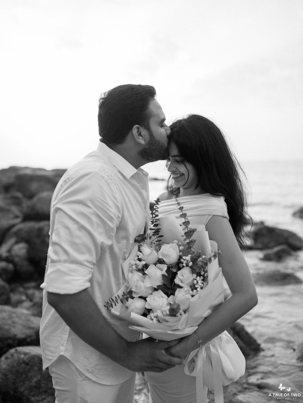 Black and white portrait of romantic couple embracing with bouquet by the ocean.
