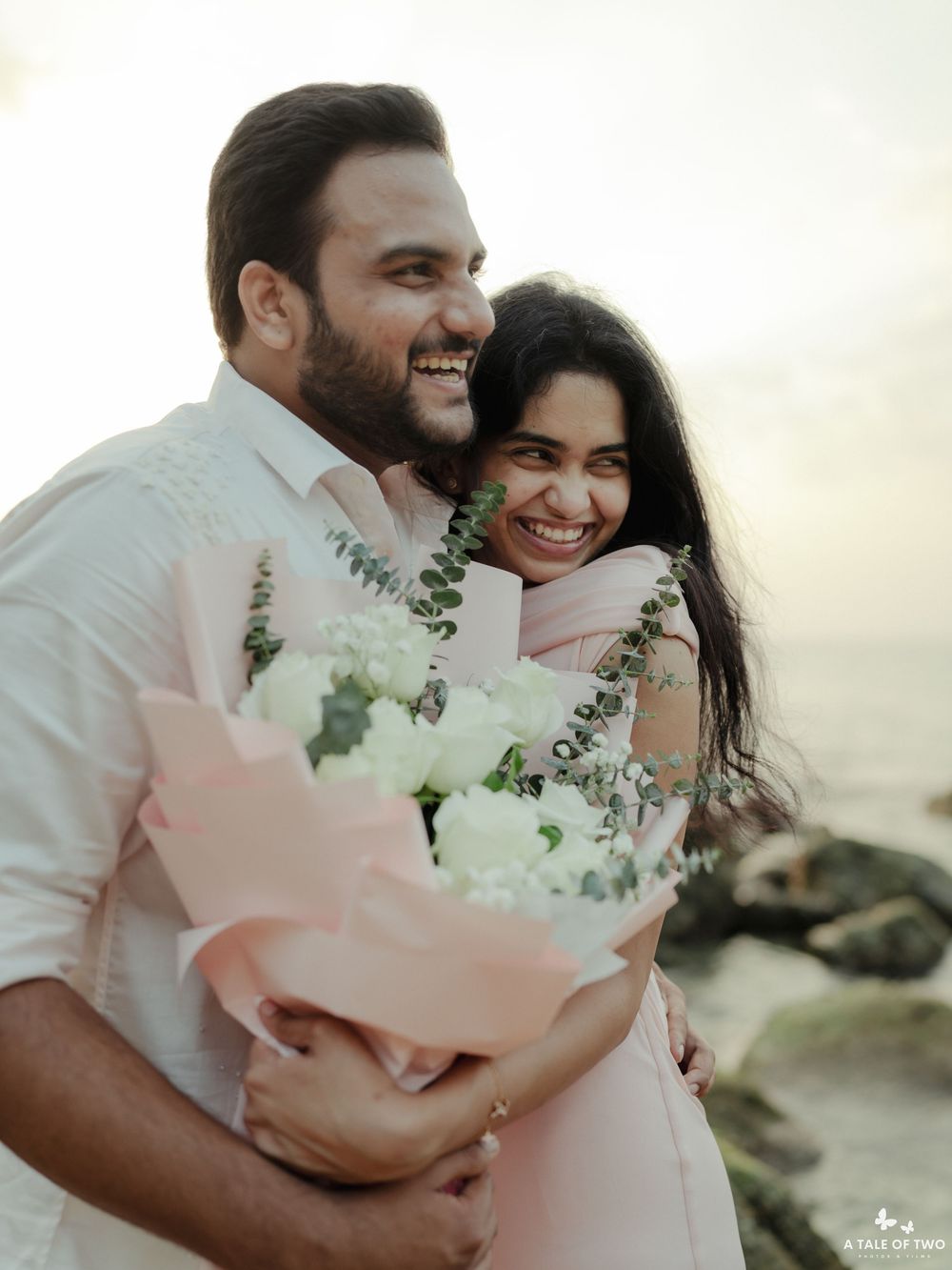 A couple in white and pink clothing holding a bouquet of white flowers on a rocky beach.