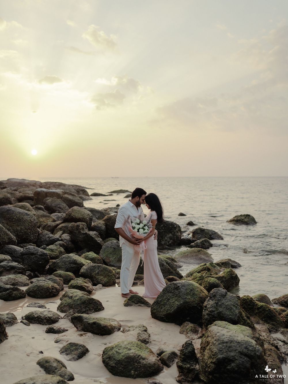 A romantic sunset beach photoshoot with a couple embracing among rocky coastal formations.