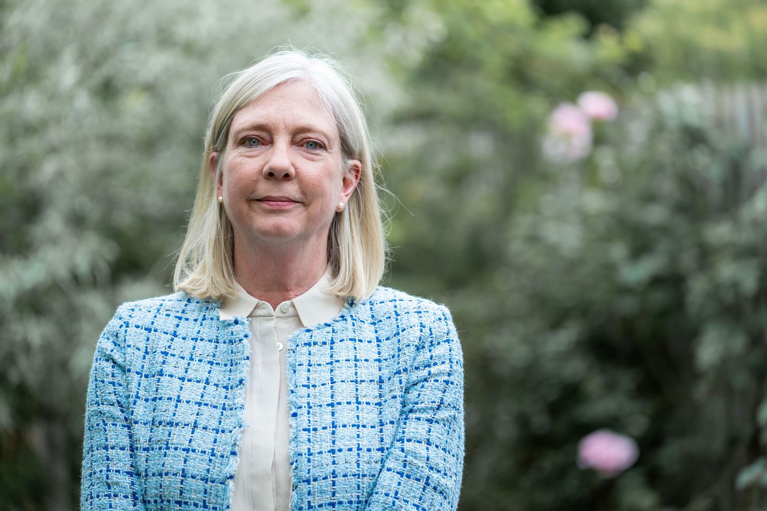 Woman in blue patterned blazer poses outdoors against green foliage background.