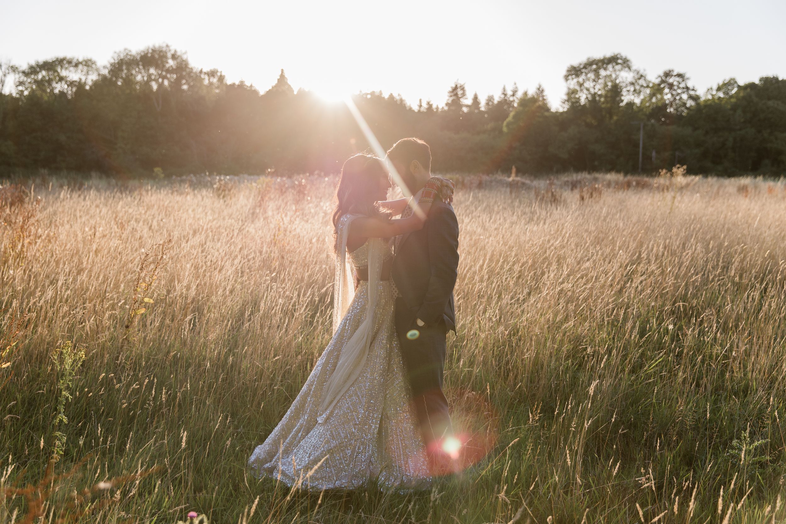 Haneef and Puja embrace in a golden sunlit meadow with tall grass at sunset, photographed by Maddie Waters at Northbrook Park