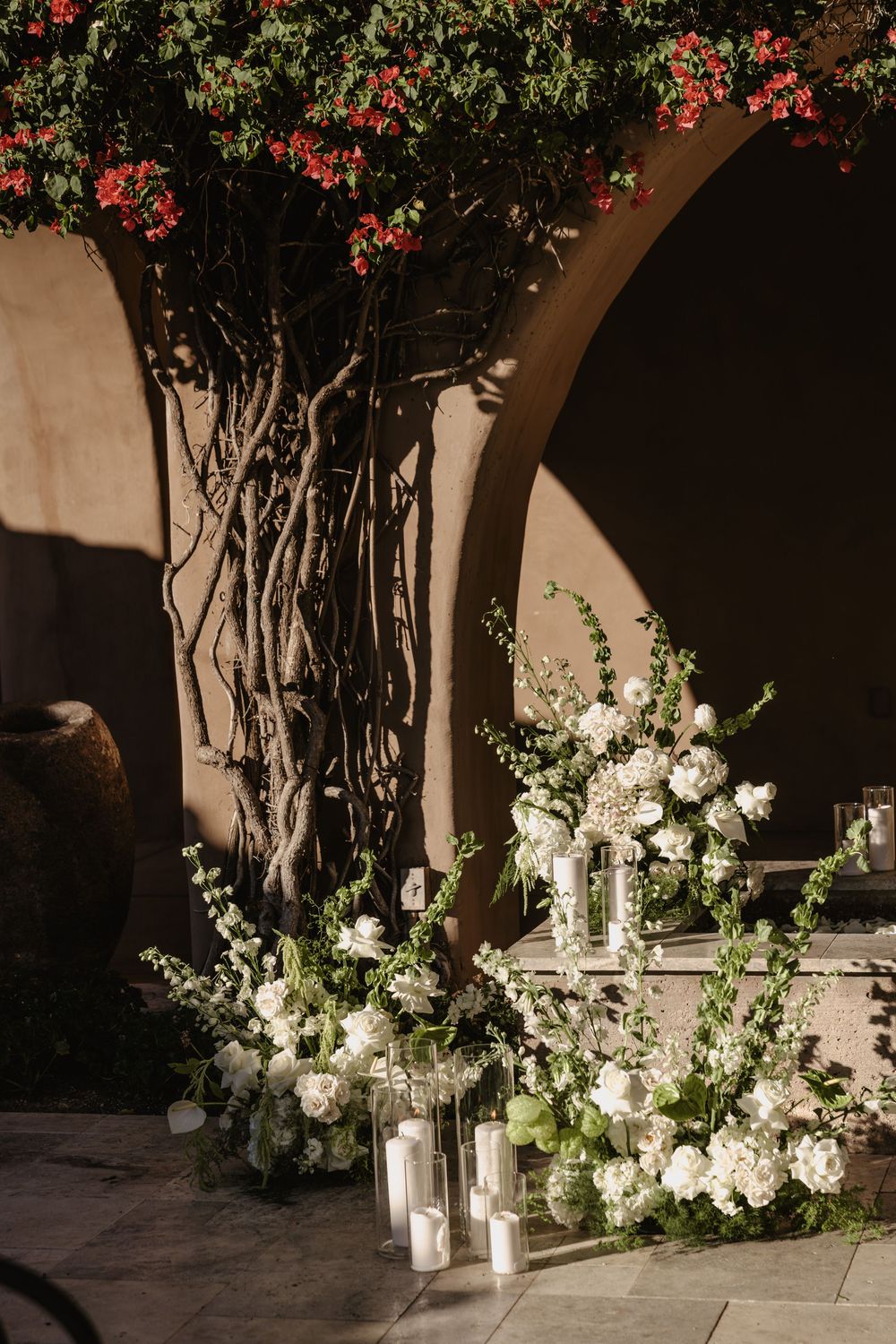 Elegant white floral arrangements with lanterns sit beneath a flowering vine-covered archway in a Mediterranean courtyard setting.