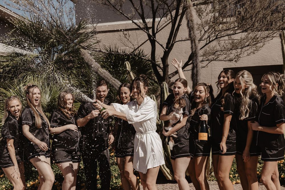 A group celebrates outdoors with champagne spray while wearing black and white outfits in the sunlight.