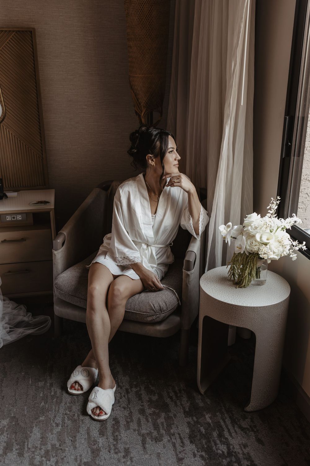 A person in a white silk robe sits thoughtfully by a window with white flowers on a side table in a moody hotel room.
