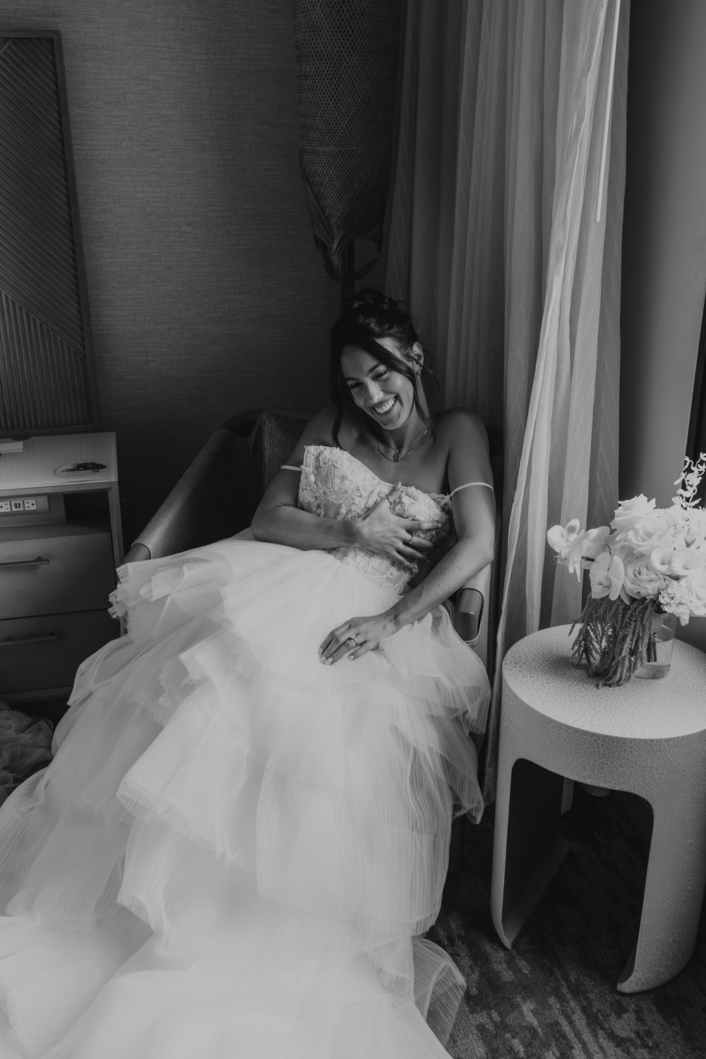 Black and white photo of someone in a flowing white wedding gown reclining on a couch beside floral arrangement.