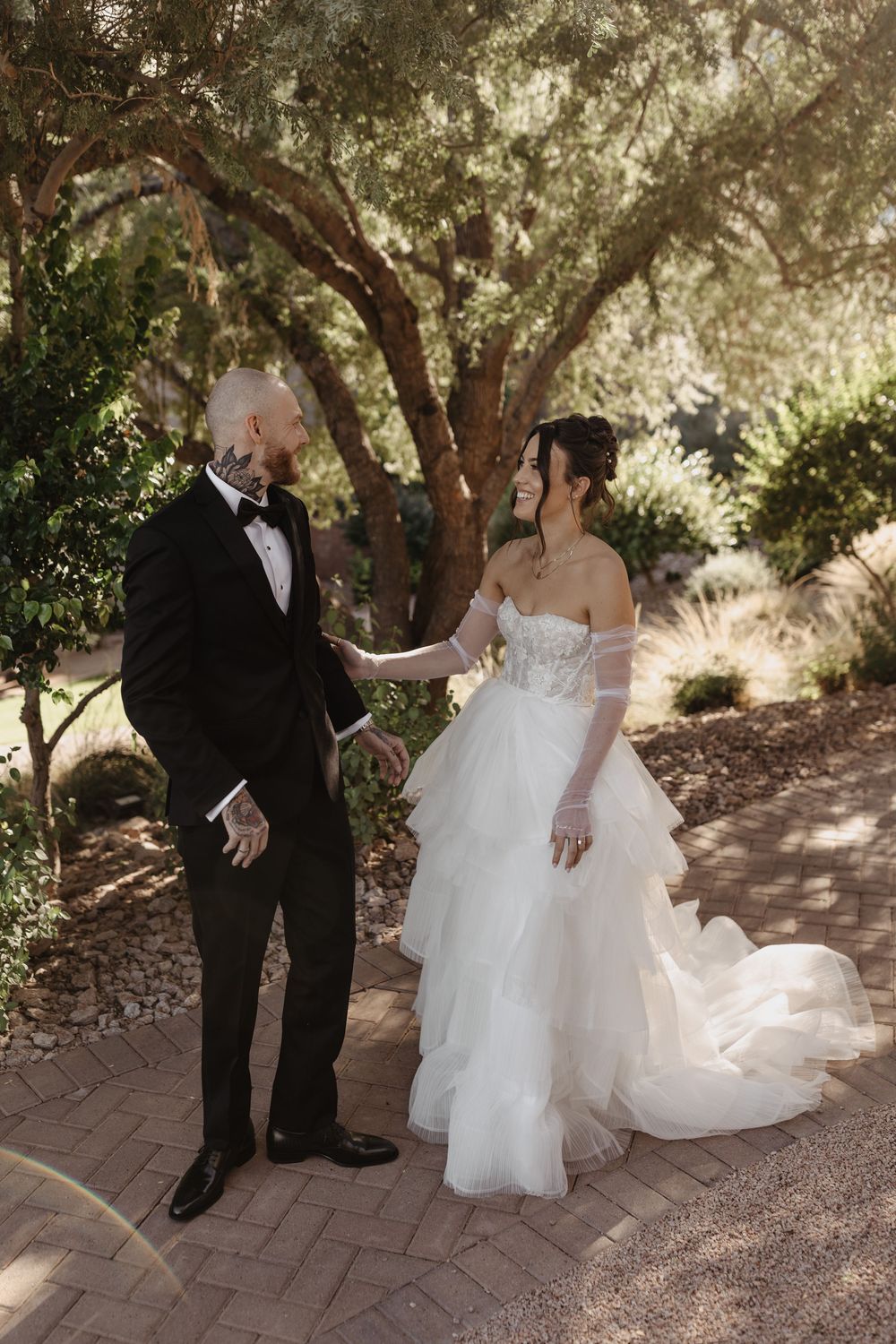 A romantic wedding couple stands together on a brick pathway under the dappled shade of garden trees.