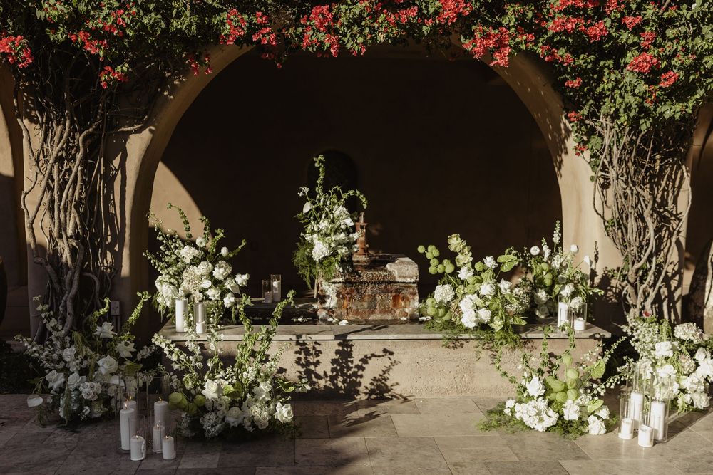 Romantic wedding altar decorated with white flowers and red bougainvillea under an elegant stone archway.