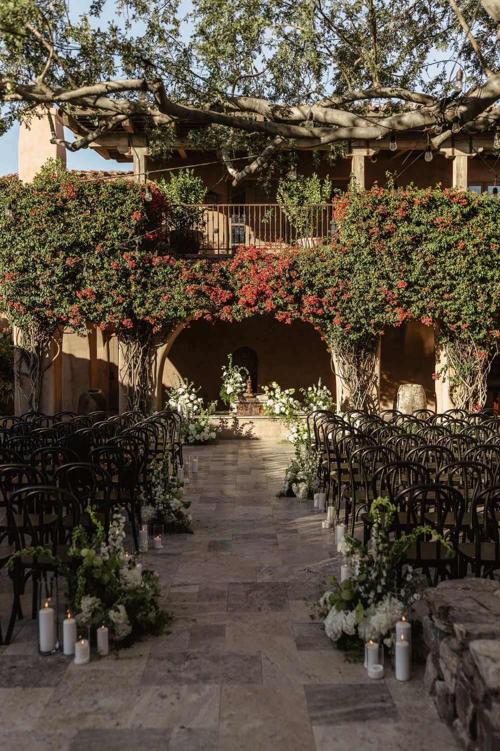 A Spanish-style outdoor wedding venue with climbing vines, white candles, and black chairs lining a stone pathway.