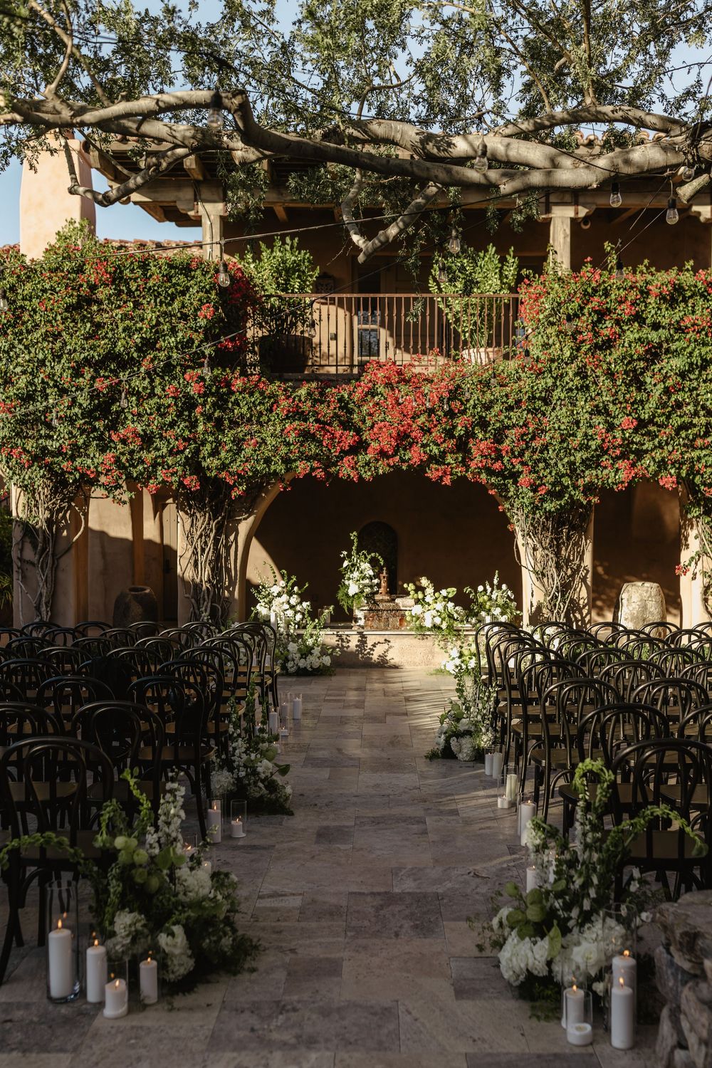 Outdoor courtyard with stone columns, flowering red bushes, and black chairs arranged in rows for an event setting.