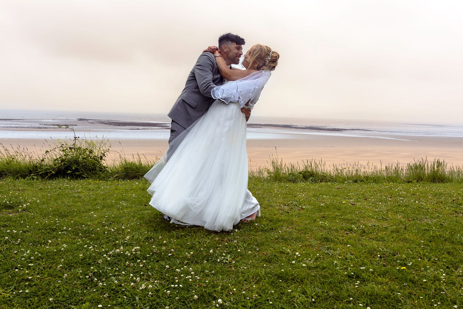 A newlywed couple shares a romantic kiss on a grassy coastal clifftop, with a stunning beach and ocean backdrop.
