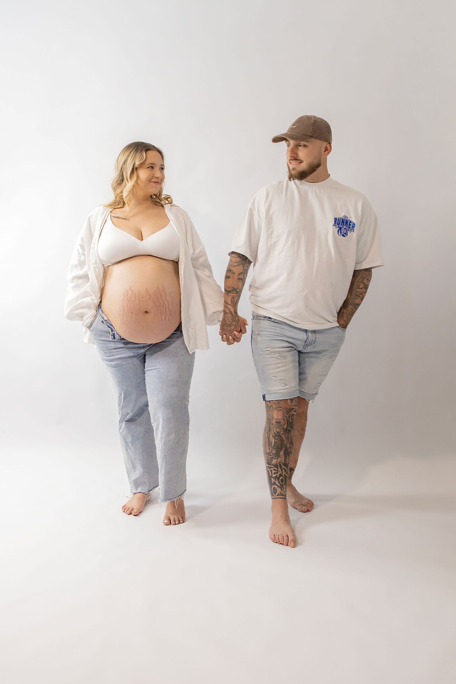 Pregnant couple holding hands barefoot on white background showing baby bump.