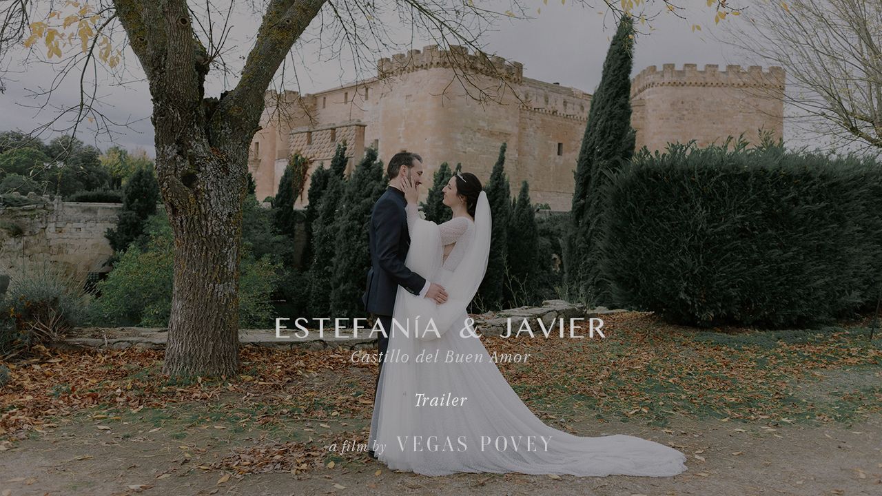 A couple in wedding attire embraces in front of a stone castle with cypress trees and autumn foliage.