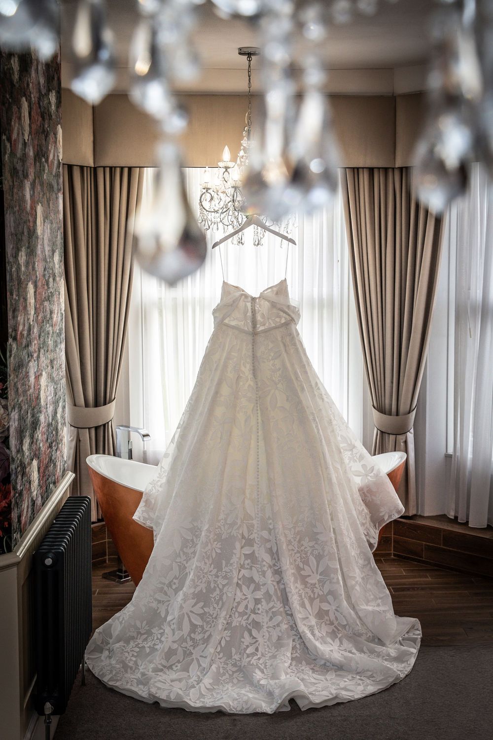 Stunning lace A-line wedding dress hanging on decorative hanger in front of window in bridal suite.