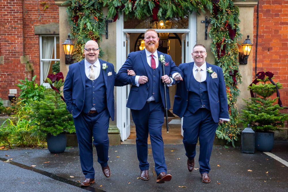 Three groomsmen in navy blue suits walking confidently outside decorated wedding venue entrance.