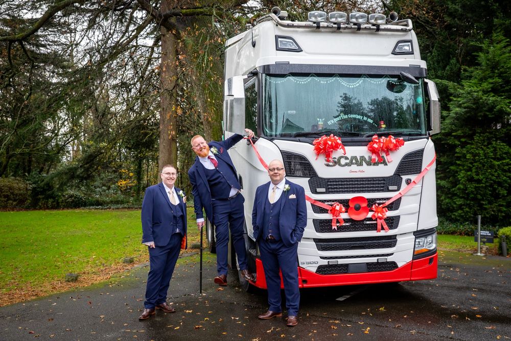 Three men in suits posing proudly in front of decorated Scania truck with red bows outdoors.