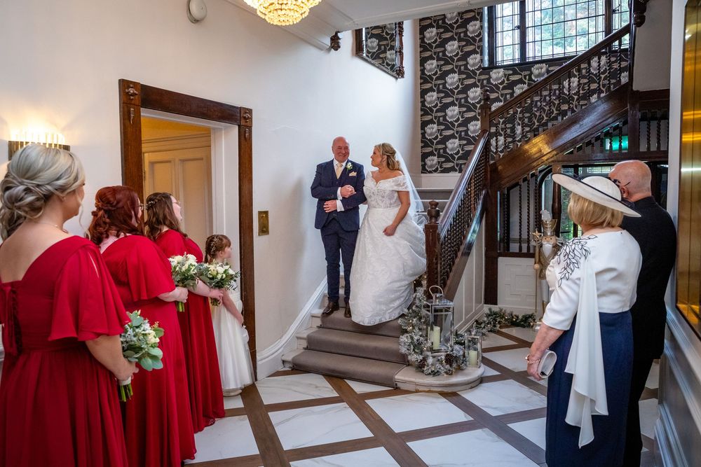 Bride descending grand staircase with father past red-dressed bridesmaids at elegant wedding venue.