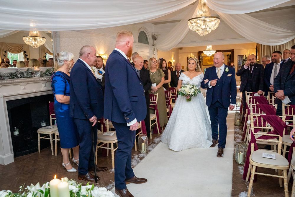 Bride walking down the aisle with father toward groom at elegant chandelier-lit wedding ceremony.