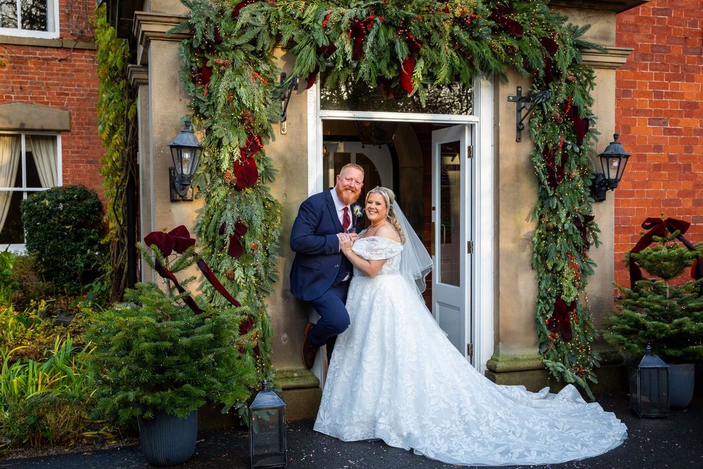 Bride and groom pose under festive Christmas floral arch at elegant brick manor house wedding venue.