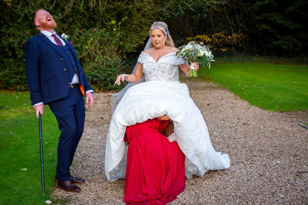 Bridesmaid in red dress adjusts bride's gown while groom laughs on garden path at wedding.