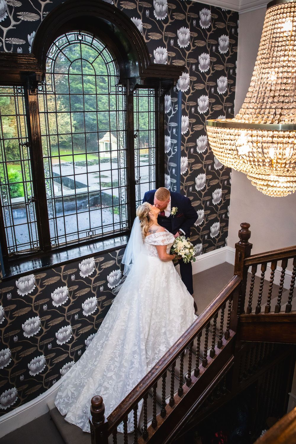 Bride and groom share romantic kiss on grand staircase with crystal chandelier and floral wallpaper.