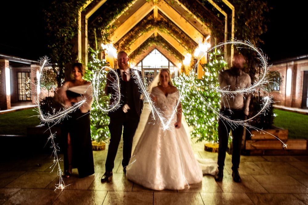 Wedding party creates sparkler LOVE sign at night outside illuminated rustic venue entrance.