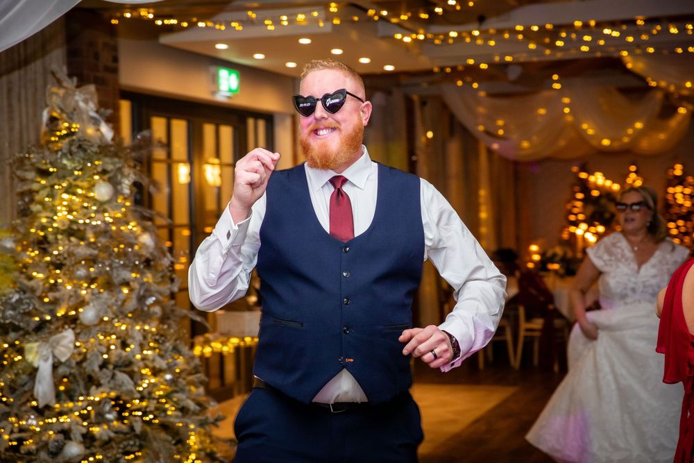 Red-bearded groomsman dances confidently in navy waistcoat and sunglasses at festive wedding reception.