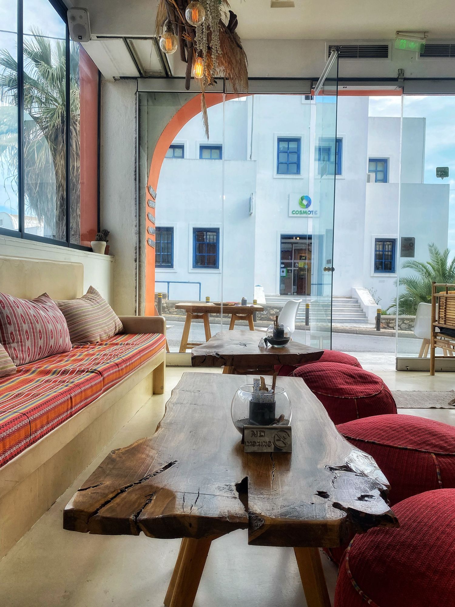 A rustic wooden live edge table with red cushioned seating in a bright modern space with palm trees visible outside.