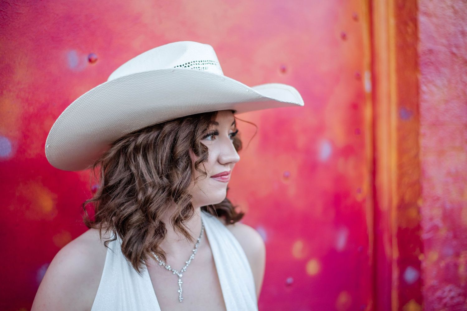 Young woman in white cowboy hat and halter dress poses against vibrant red and pink wall.