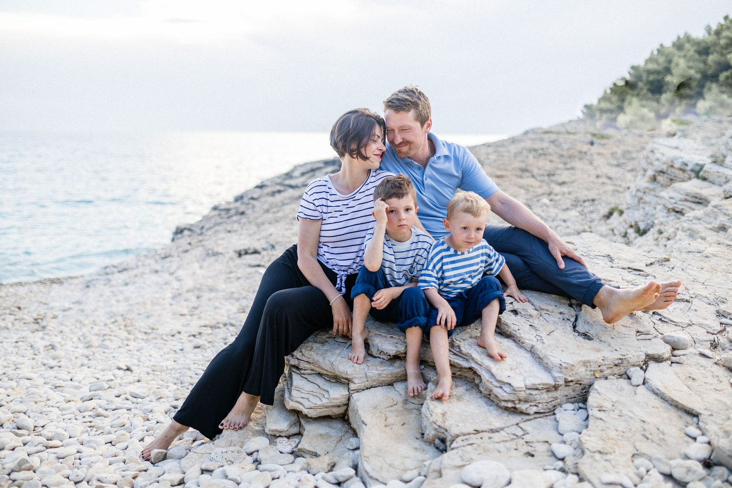 A family in coordinating blue and white outfits sits together on coastal rocks by the ocean.