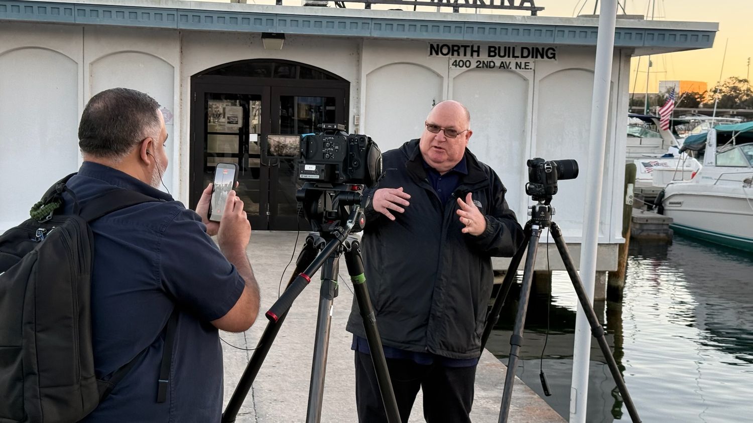 Cameraman filming a man being interviewed outdoors near a marina with tripod-mounted cameras at dusk.