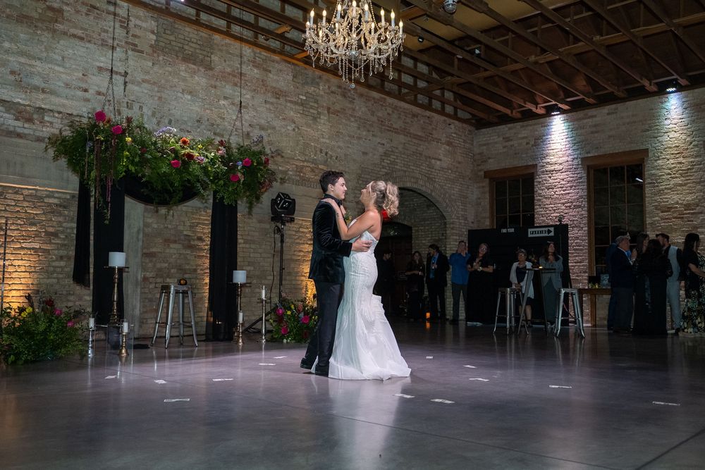 A couple shares their first dance in an elegant rustic venue with exposed brick walls and a crystal chandelier overhead.
