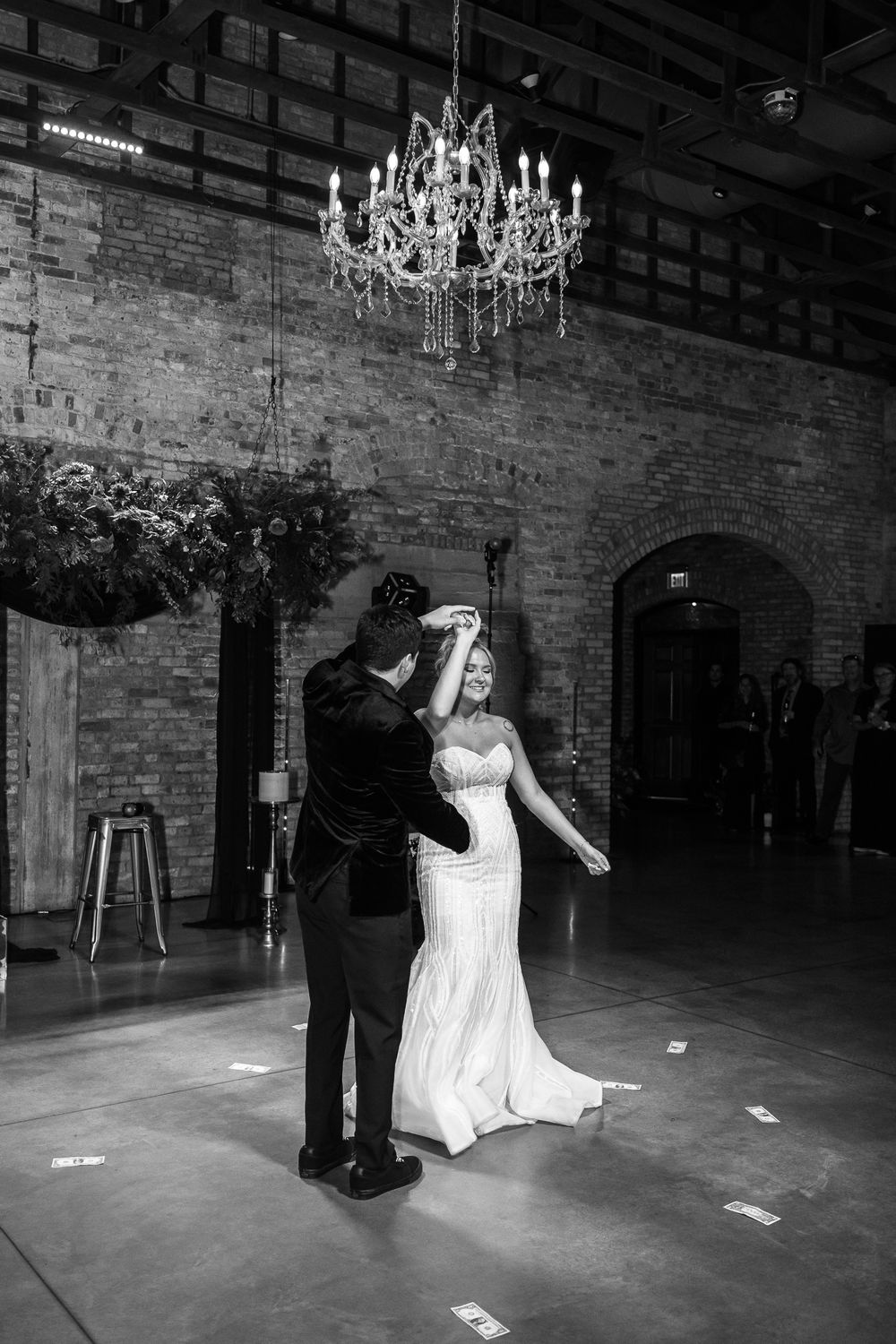 Wedding party dancing in a rustic industrial venue with exposed brick walls and elegant chandeliers overhead.