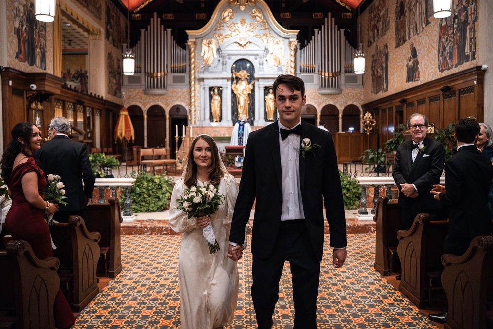 A wedding ceremony procession walking down the aisle of an ornate cathedral with gothic architecture and stained glass windows.