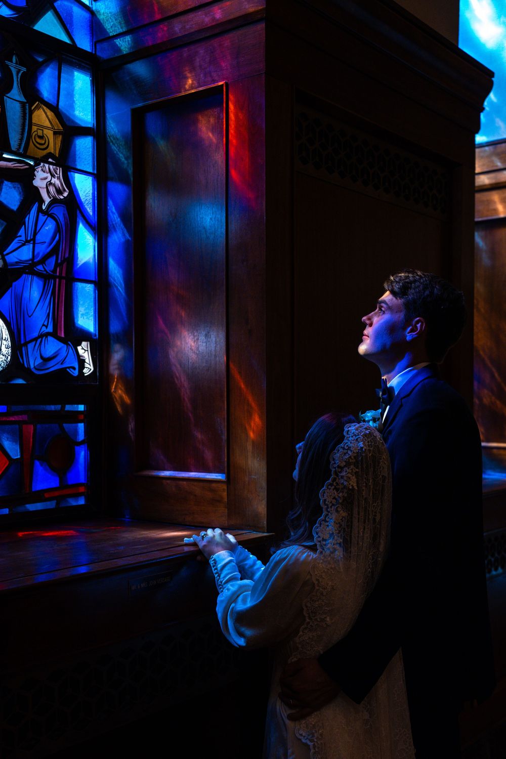 Dramatic blue lighting illuminates a figure seated in a dark room looking out a window with stained glass elements.