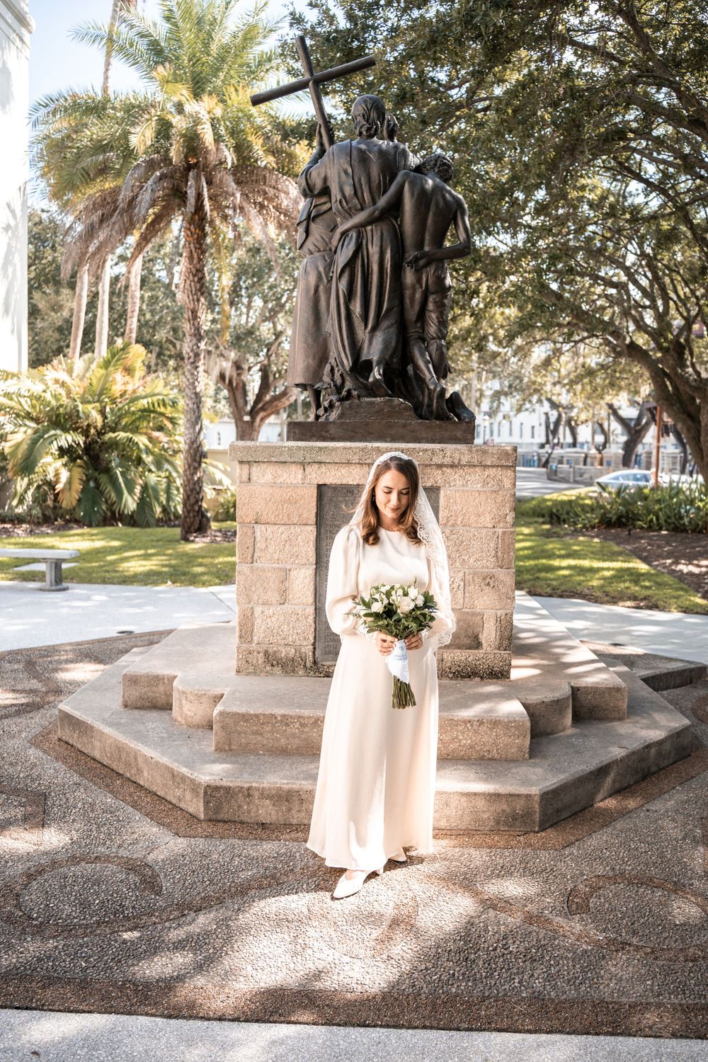 A person in a light-colored dress poses at a bronze statue monument in a park setting with palm trees and greenery.