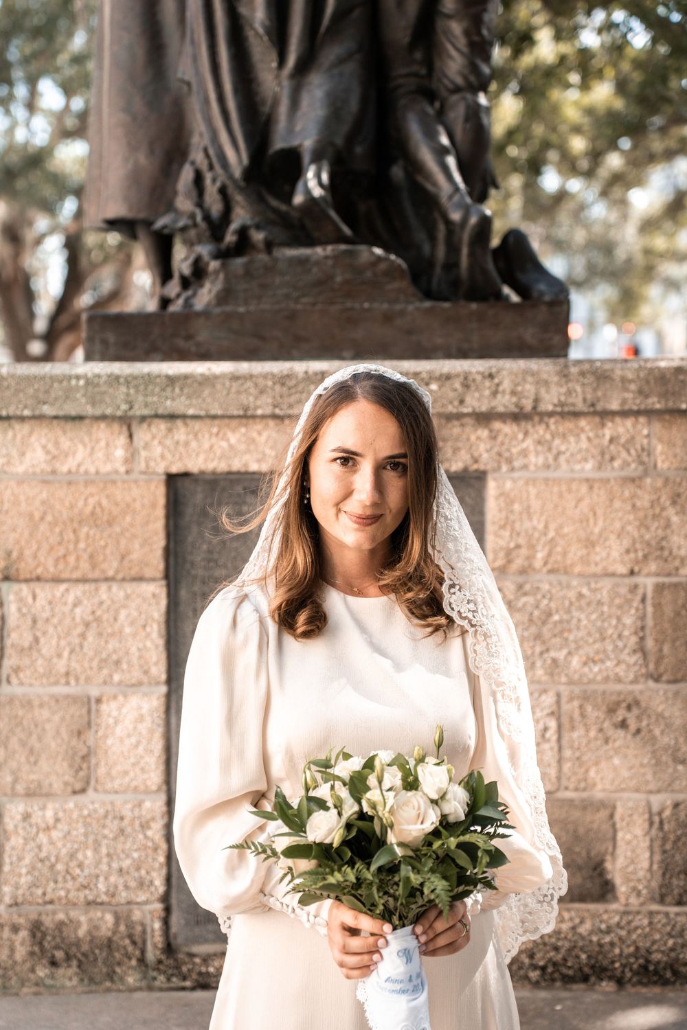 A person in a white dress holds a bouquet of white roses while standing against a stone wall outdoors.